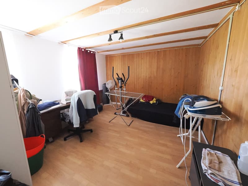 A small, furnished room with wooden flooring and slanted wooden ceiling beams. Contains a desk with a chair, an ironing board with an iron, a fan, an exercise bike, and a curtain on the window.