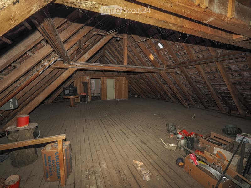 An attic space with exposed wooden beams and rafters, wooden flooring, and minimal furnishings. Visible items include a small table, a few boxes, and scattered tools.