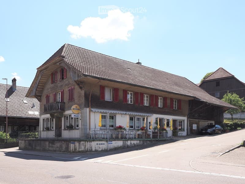 Two-story building with red shutters, a balcony, and outdoor seating area with tables and chairs.