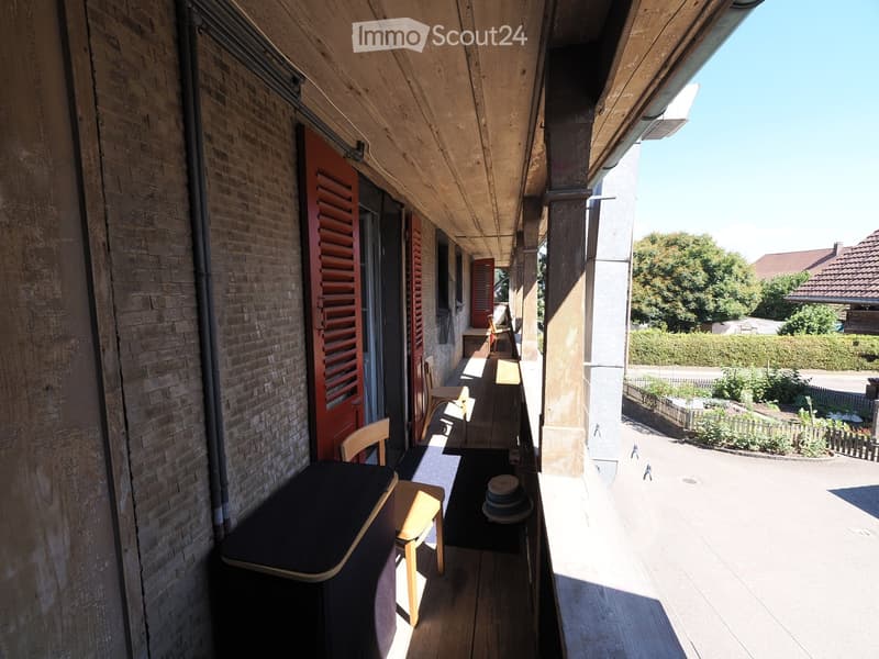 Balcony with wooden floor, table, chairs, and a view of the street and greenery.