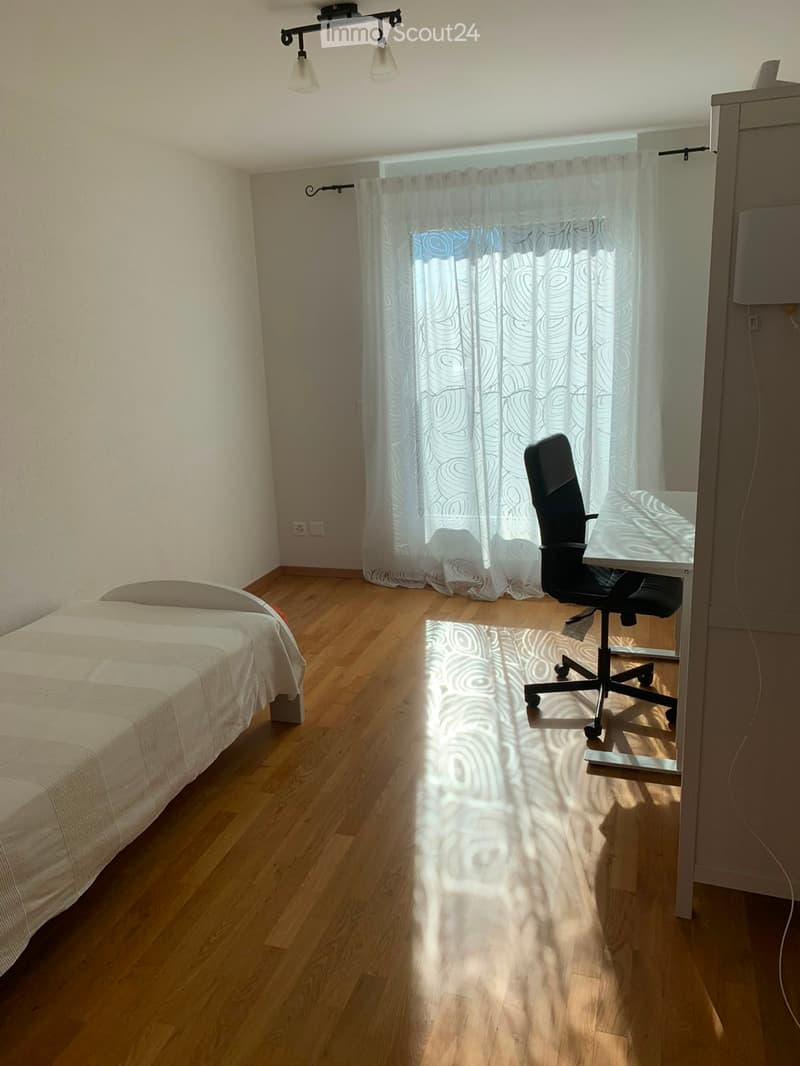 Bright bedroom with wooden flooring, a single bed with white bedding, a desk with a chair, and a window with a patterned curtain.