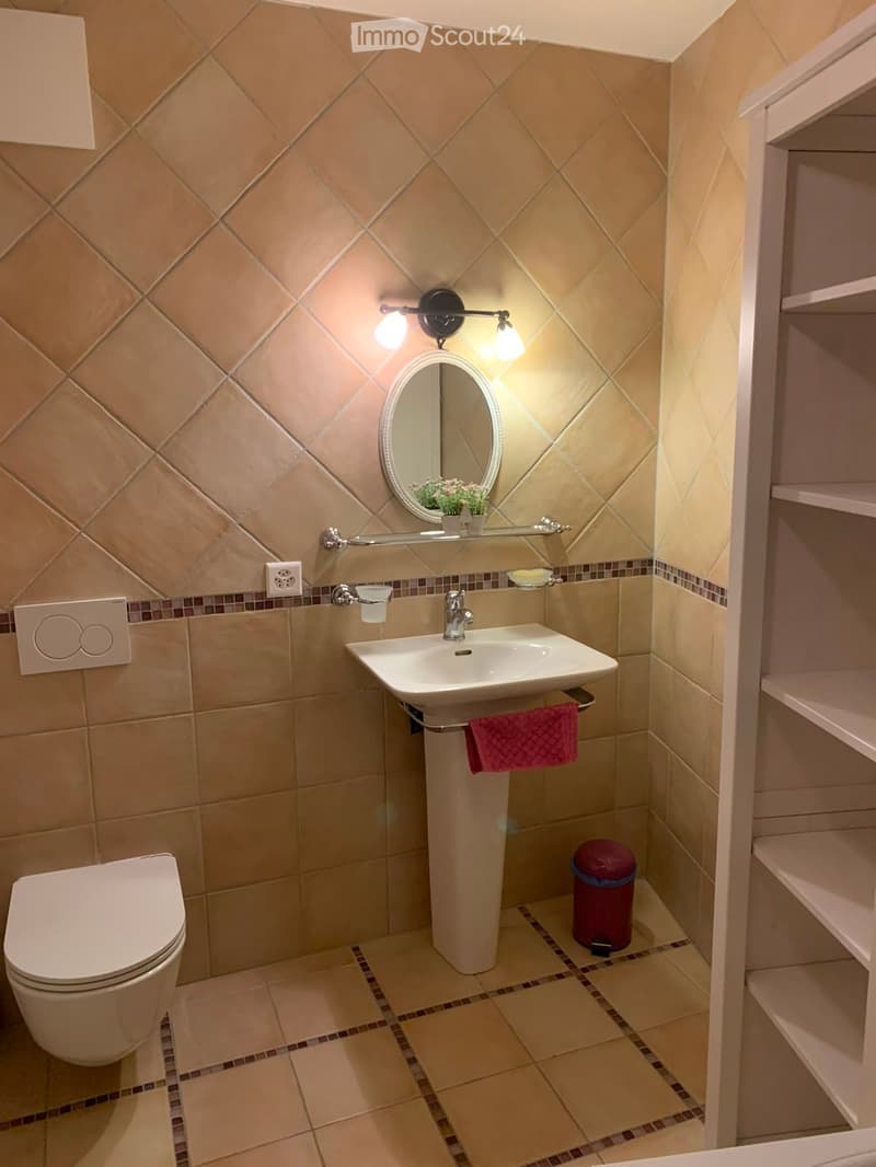 Modern bathroom with beige tiles, a white toilet, a sink with a mirror above it, a towel hanging on the side, and a small shelf with decorative items.