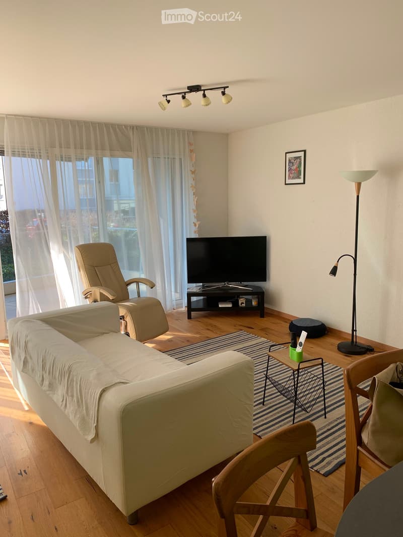 Bright living room with wooden flooring, large windows with sheer curtains, a beige sofa, a matching armchair, a coffee table, a television on a stand, and a floor lamp.