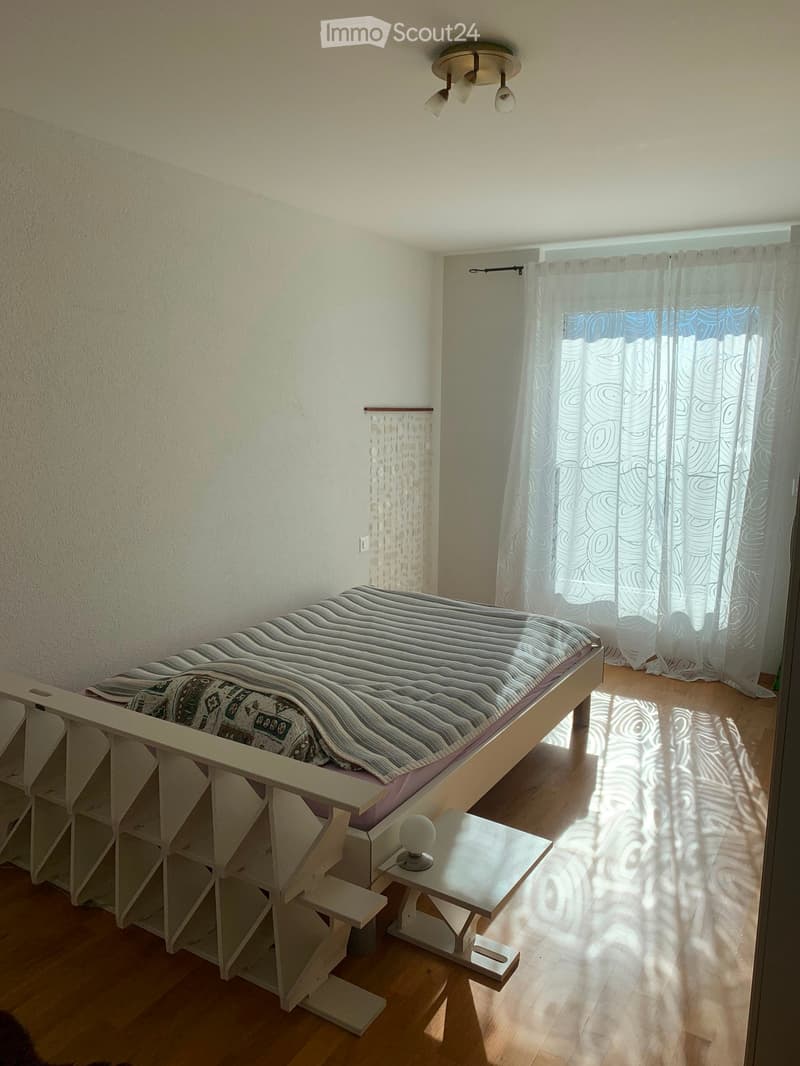 A small, well-lit bedroom with a wooden floor, a bed with striped bedding, a nightstand, a small table, and a chair. The room has white walls and large windows with sheer curtains.