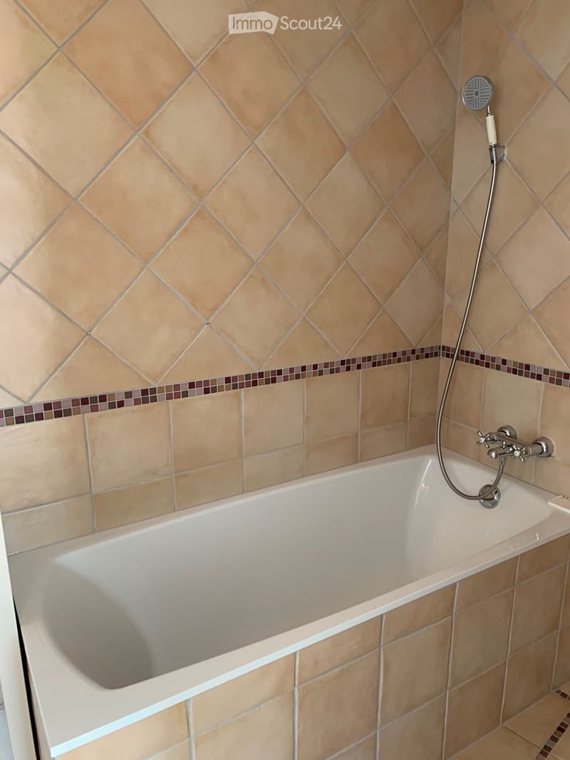Bathroom with a white bathtub, beige tiled walls, and a showerhead attached to the wall.