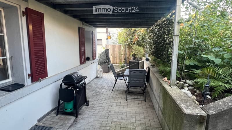 Balcony with brick floor, barbecue, chairs, and table, surrounded by greenery and a wooden fence.