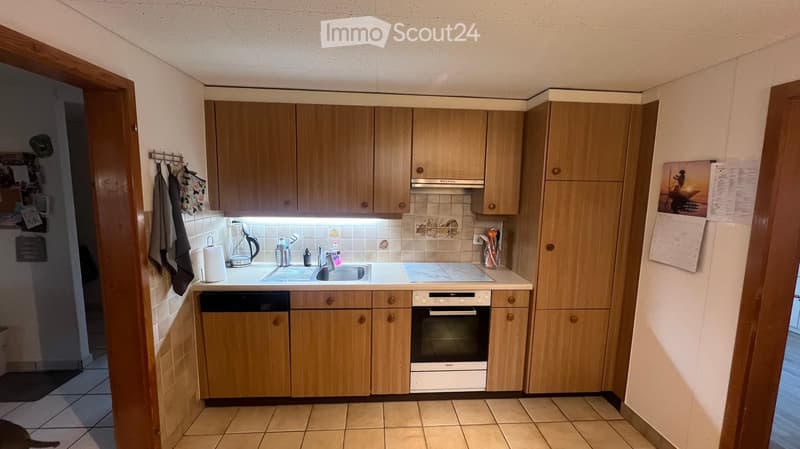 A compact kitchen with light wooden cabinets, a white countertop, and built-in appliances including a sink, oven, and dishwasher. The space features tiled flooring and walls, with a small window above the sink.