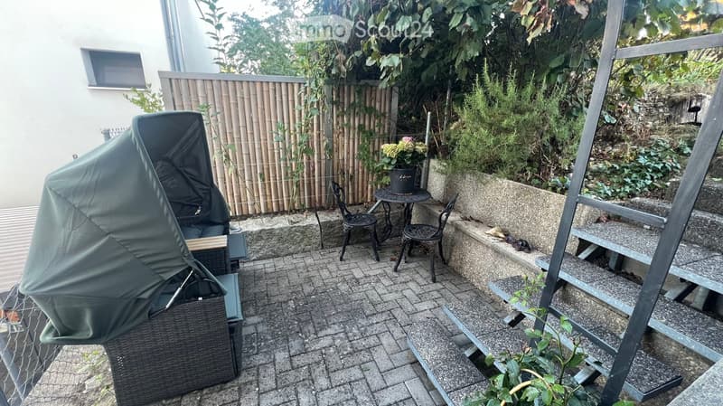 Small outdoor patio with a covered umbrella, a small table, and two chairs, surrounded by greenery and a bamboo fence.