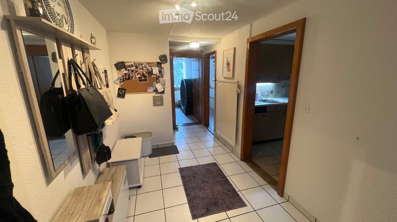 A well-lit hallway with tiled flooring, wooden doors, and a coat rack with several bags. A bulletin board with photos and notes is visible on the wall, and a small table and trash bin are placed near the entrance to another room.