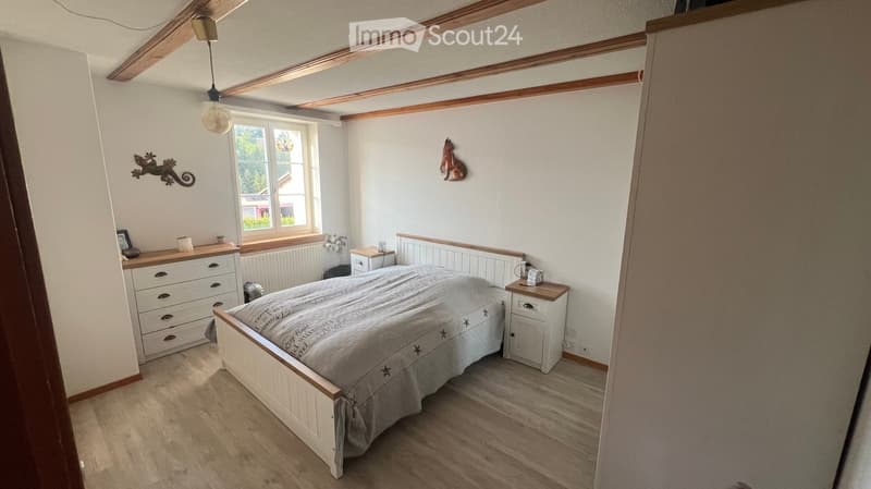 Cozy bedroom with wooden flooring, white walls, and exposed wooden beams on the ceiling. Features a bed with a grey duvet, a nightstand, a drawer unit, and wall-mounted decorative items.