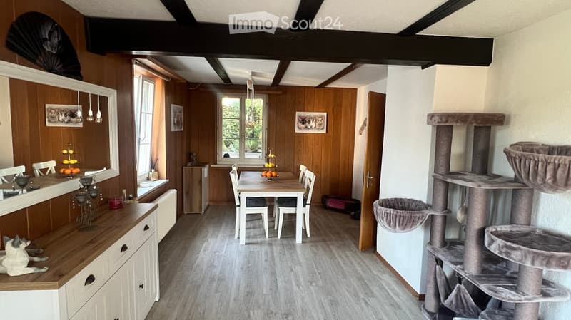 A cozy, well-lit room with wooden walls and a high ceiling. Features include a dining table with chairs, a cat tree, a dog resting on a counter, and decorative fruit bowls on the counter.