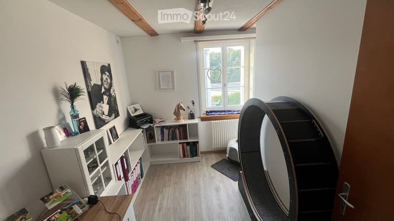 A cozy, well-lit room with wooden flooring, white walls, and slanted wooden ceiling beams. Features include a large window with a heart-shaped cutout, a white shelving unit with books and decorative items, a small table with a vase and picture frames, and