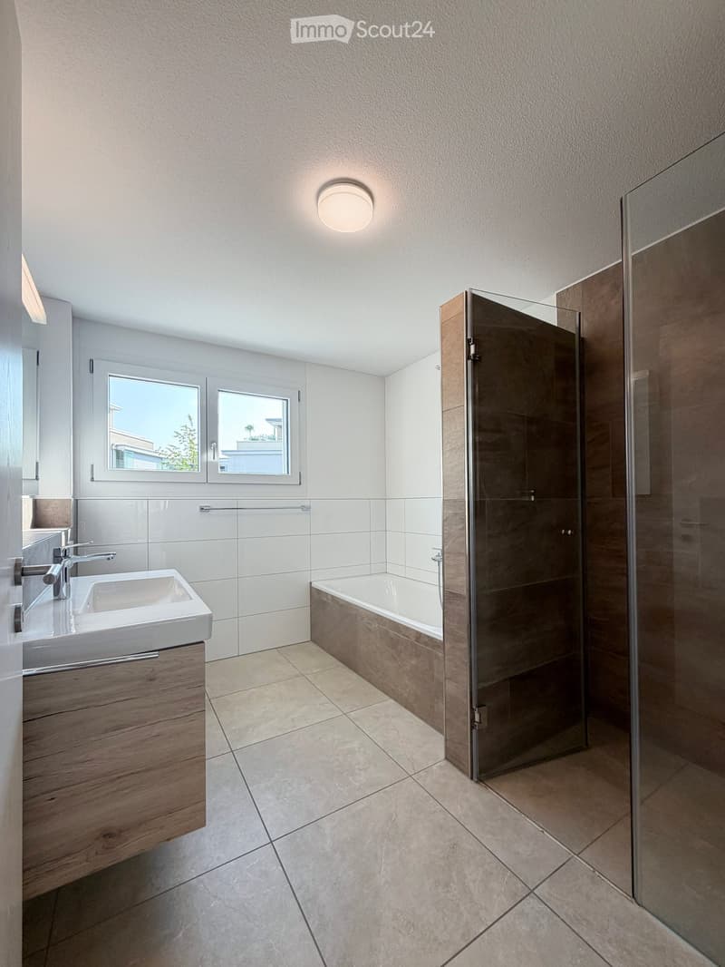 Modern bathroom with a white tiled wall, a wooden vanity with a sink, a bathtub, and a glass-enclosed shower. The room features large windows offering a view of the outdoors.