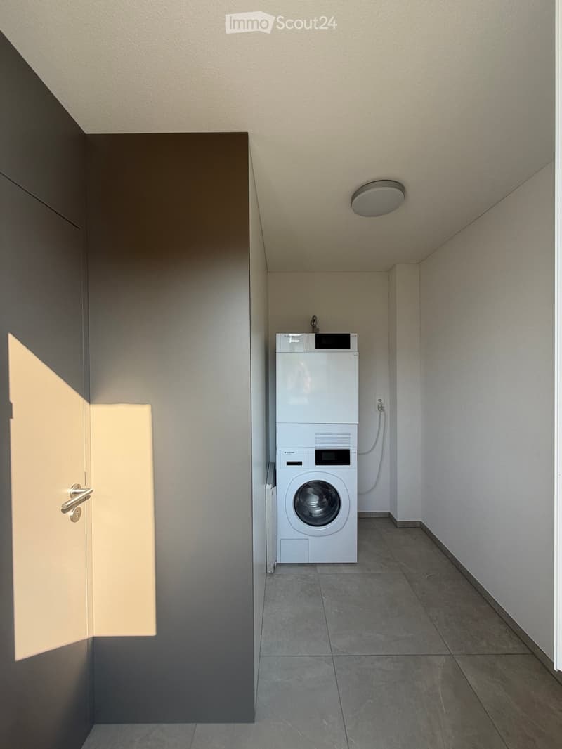 A narrow hallway with a white washing machine and dryer unit against one wall, a door on the left, and a ceiling light fixture.