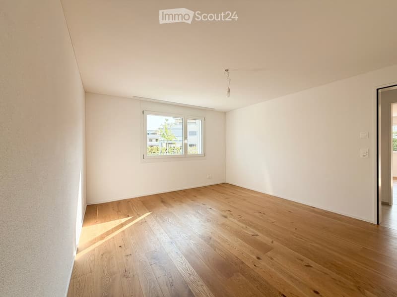 Empty room with light wooden flooring and white walls, featuring a window with white frames and a visible light fixture on the ceiling.