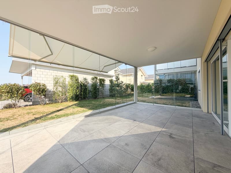 Modern outdoor space with large glass walls, tiled floor, and a retractable awning, overlooking a landscaped garden.