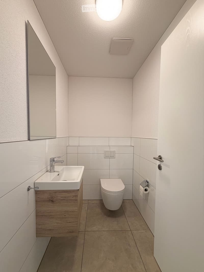 Modern bathroom with a white sink, chrome faucet, toilet, and a mirror on the wall. The room features light-colored tiles and a wooden cabinet under the sink.