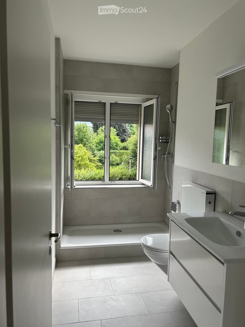 Modern bathroom with a bathtub, toilet, sink, and mirror. Large window with blinds offers a view of greenery outside.