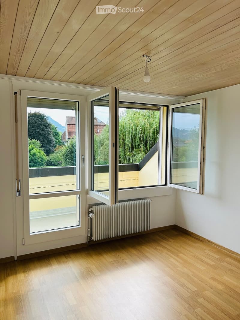 Empty room with wooden flooring, white walls, large windows with views of greenery and mountains, and a radiator under one window.