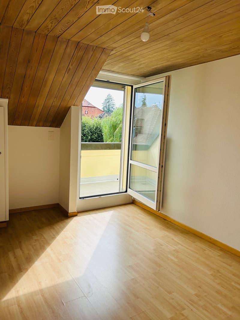 Bright, empty attic room with wooden floors, slanted wooden ceiling, and large windows offering natural light and a view of the outdoors.
