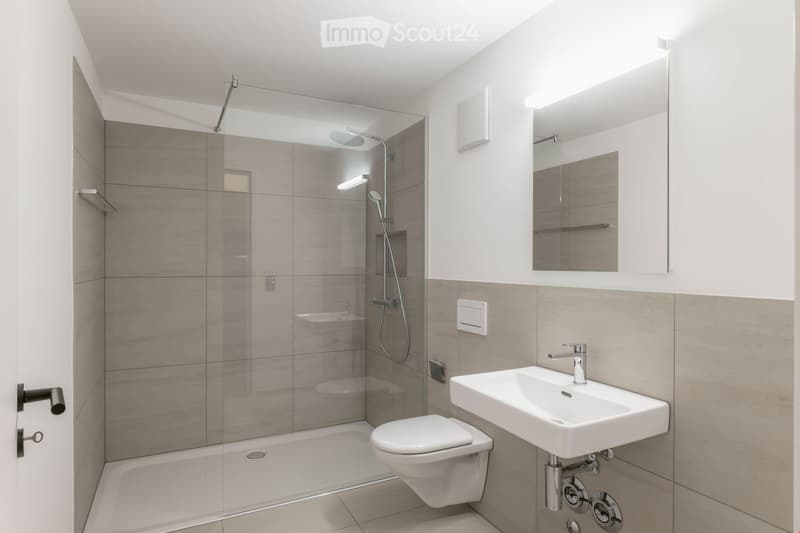 Modern bathroom with a glass-enclosed shower, bathtub, toilet, sink, and mirror. The walls are tiled in neutral tones, and the space is well-lit.