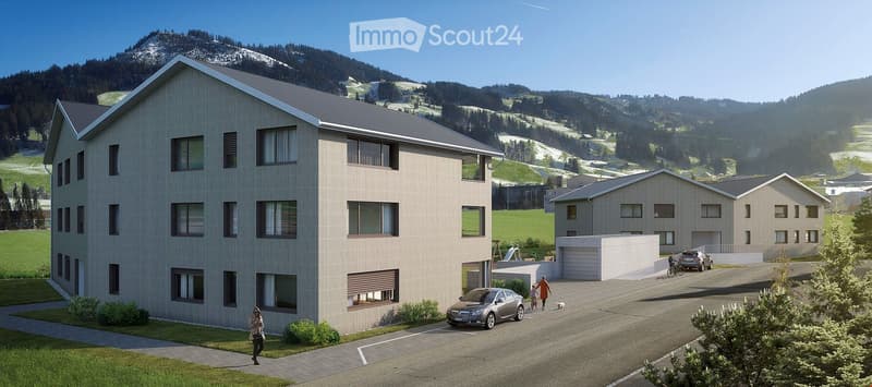 Modern multi-story residential building with large windows and a flat roof, surrounded by a parking area and greenery, with a scenic mountain backdrop.