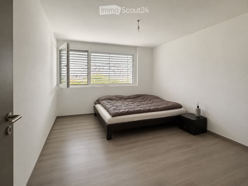 Minimalist bedroom with a single bed, nightstand, and lamp. The room features light wooden flooring and white walls, with a window offering a view of greenery outside.