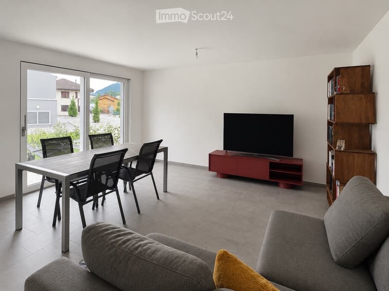 Modern living room with a grey sofa, a wooden shelf, a flat-screen TV on a red stand, a dining table with four chairs, and a large window with a view of the outdoor area.