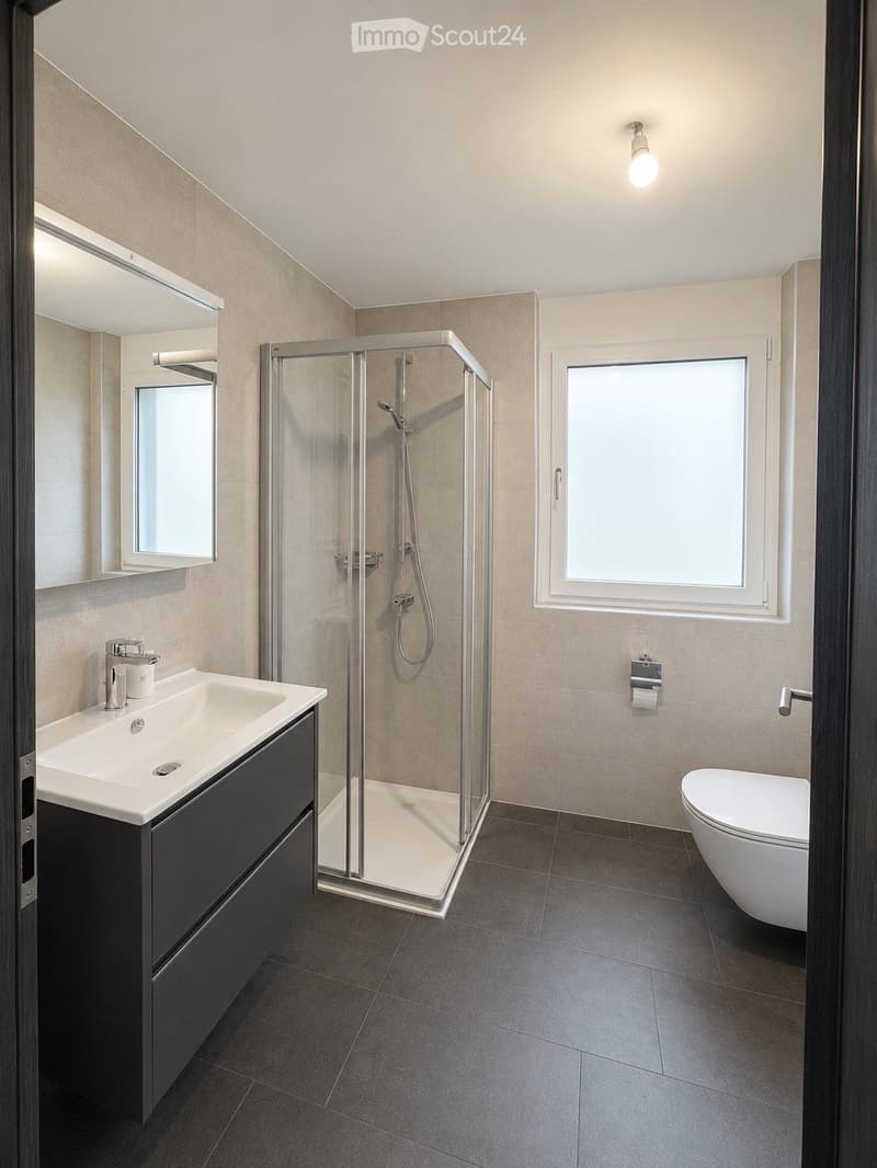 Modern bathroom with a white sink, dark vanity, large mirror, glass-enclosed shower, and toilet. The room features beige tiles and a window providing natural light.