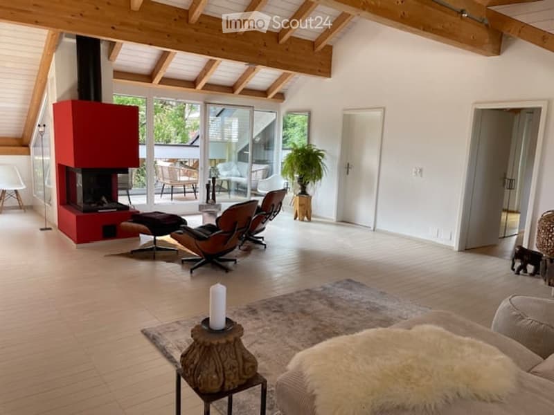 Bright living room with wooden floors, exposed wooden beams, large windows, fireplace, sofa, coffee table, ottoman, chairs, and a plant in the corner.