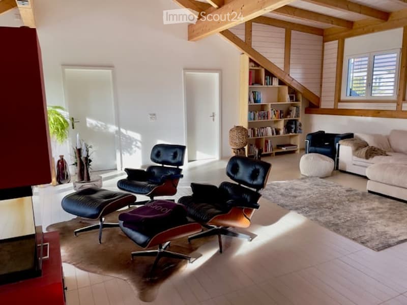 Modern living room with exposed wooden beams, white walls, and large windows. Features a cream sofa, black leather chairs, ottomans, a bookshelf, and a table lamp.