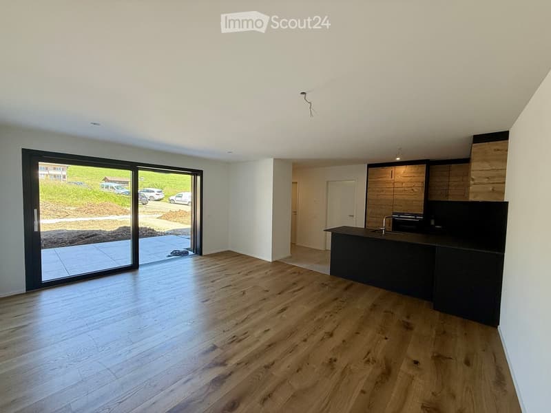 Empty room with wooden flooring, white walls, and a large sliding glass door leading to an outdoor area. A small kitchen area is visible in the background with cabinets and a countertop.