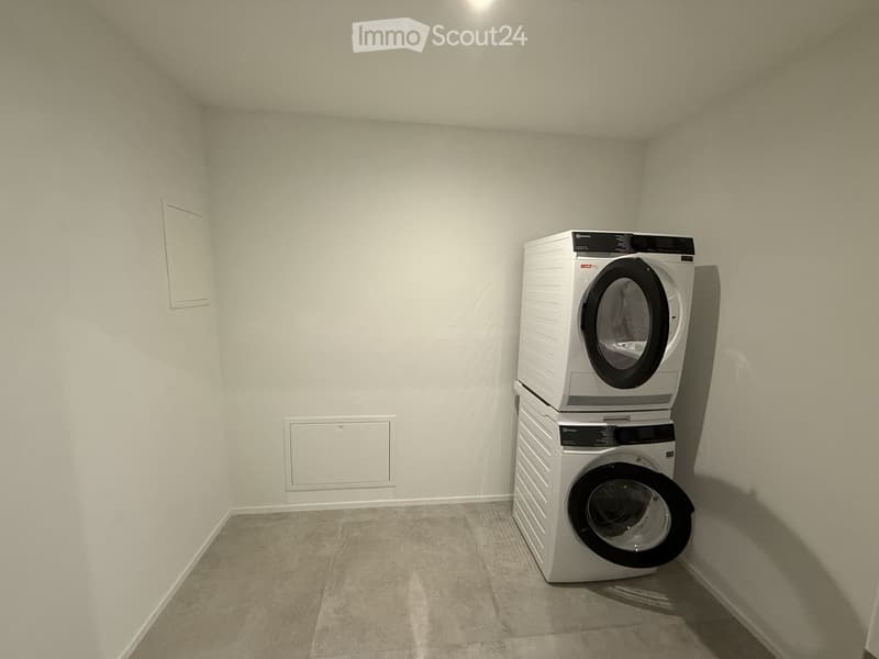 A small, unfurnished room with a white tiled floor and light grey walls, featuring a stacked washing machine and dryer unit in the corner.