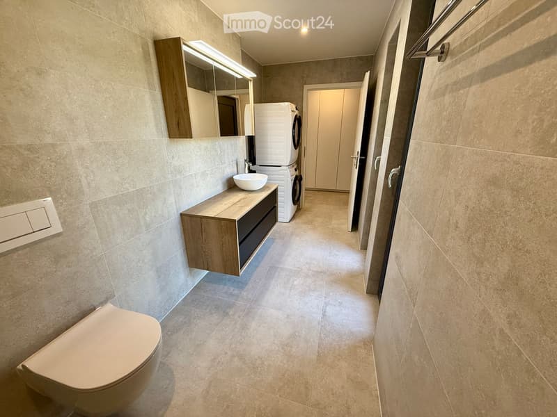 Modern bathroom with beige tiles, a toilet, a sink with a wooden cabinet, a mirror, and a washing machine in the corner.