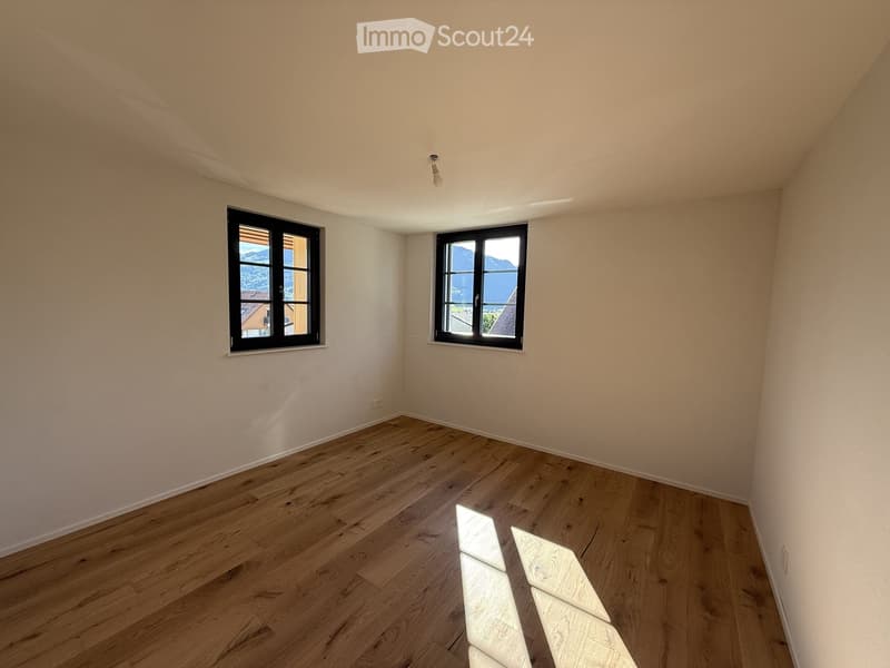 Empty room with wooden flooring and two windows offering a scenic view of mountains and a lake.