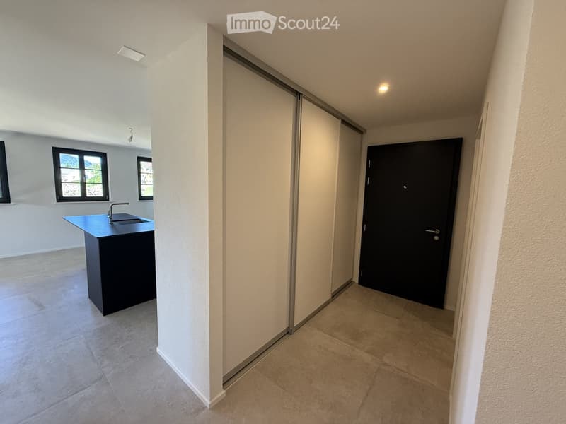 Empty hallway with tiled floors, white walls, and a black door at the end. A small kitchen area with a sink and countertop is visible to the left.