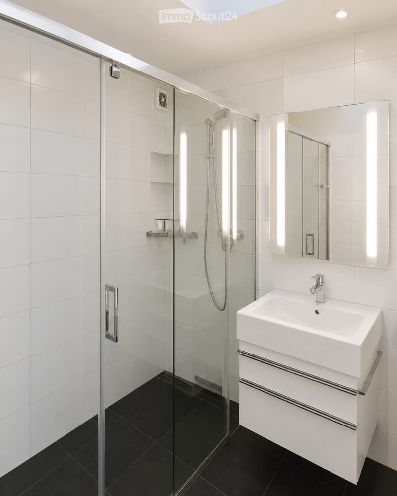 Modern bathroom with white tiles, a glass-enclosed shower, a white sink with a cabinet, and a large mirror with built-in lighting.