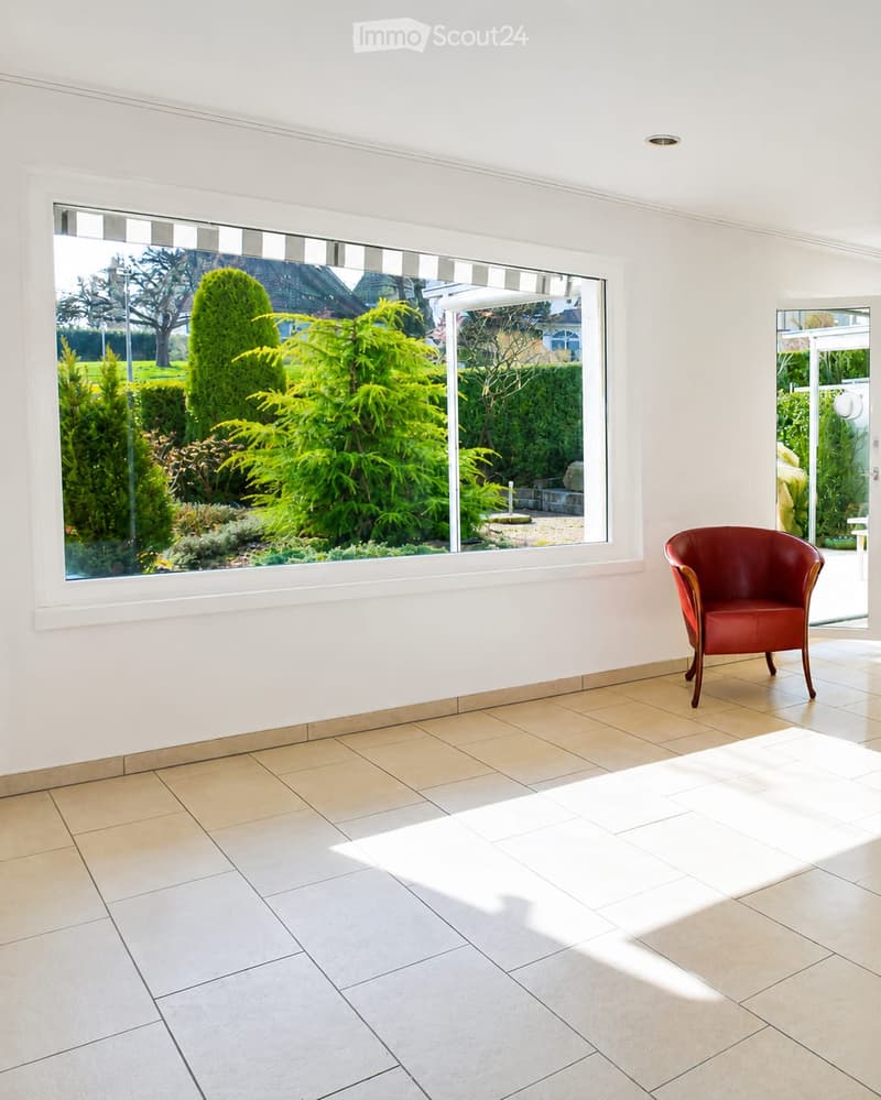 Bright, minimalist room with large windows overlooking a garden, tiled floor, and a single red leather chair.