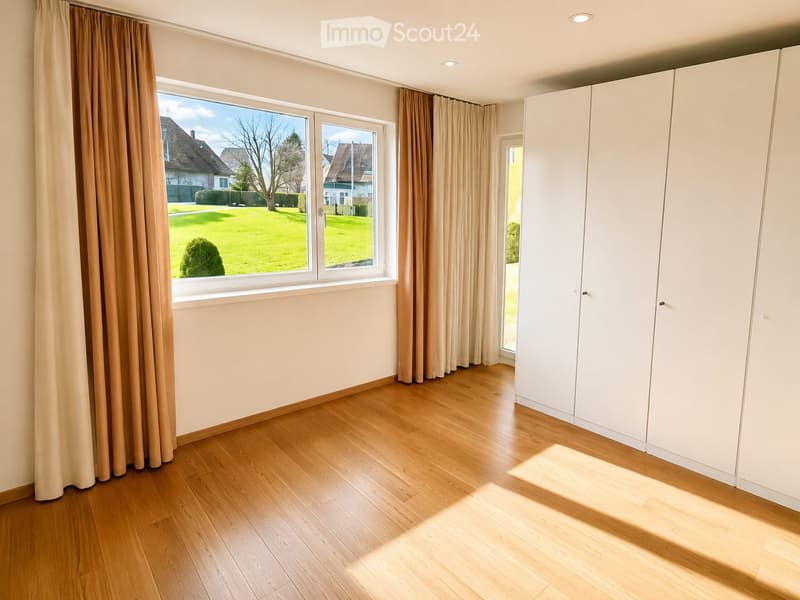 Empty room with wooden flooring, large windows with beige curtains, white built-in wardrobes, and a view of a green garden.