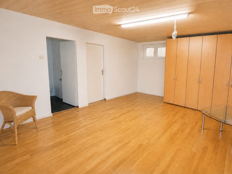 Bright, unfurnished room with wooden flooring, white walls, and a wicker chair near the wall. A glass table is positioned near a large wardrobe, and a bathroom is visible through an open doorway.