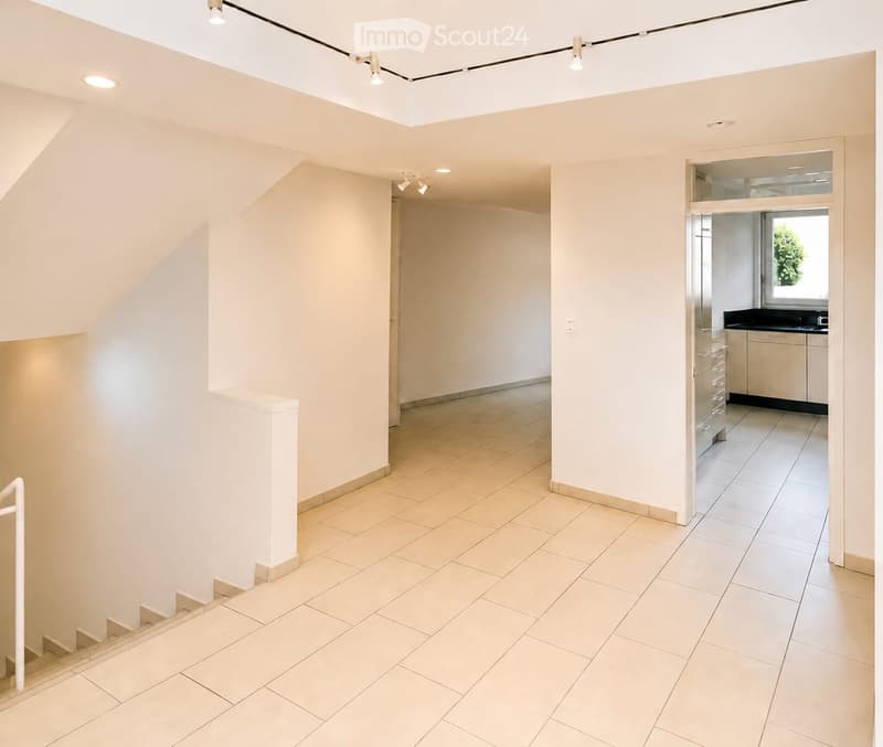 Bright, modern hallway with tiled flooring, white walls, and recessed lighting. Features a staircase leading upwards and an open doorway to a kitchen area with white cabinets and a window.