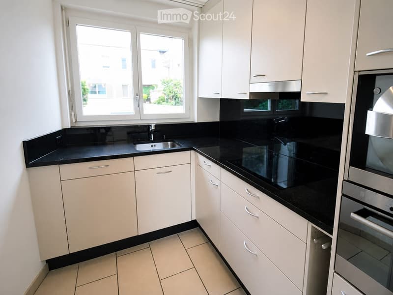 Modern kitchen with white cabinets, black countertops, and stainless steel appliances, including a sink and oven. Large windows provide natural light.