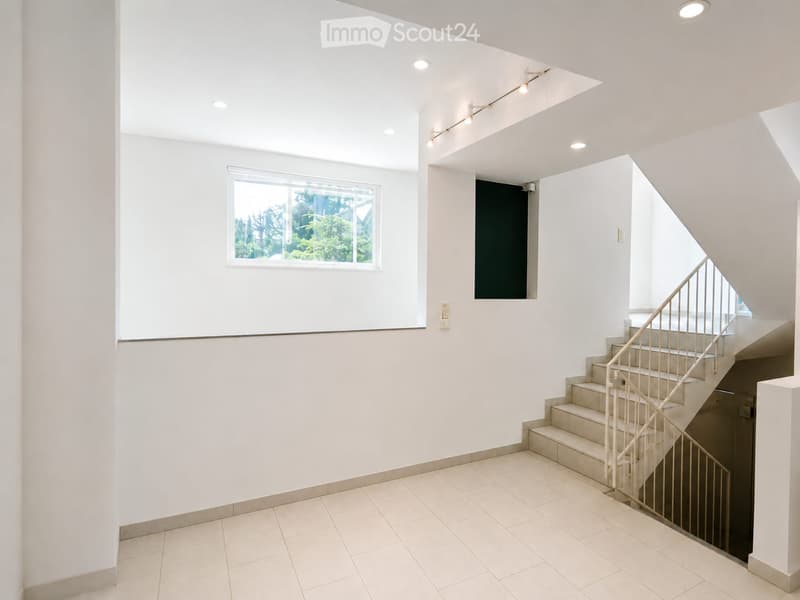 Bright, modern hallway with white walls, tiled floor, and a staircase with a metal railing leading upwards. A window on the left provides natural light, and recessed ceiling lights illuminate the space.