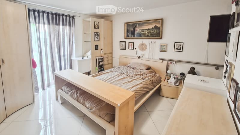 A well-lit bedroom with a large bed, nightstand, mirror, and several picture frames on the wall. The room features a curtained window and a cabinet with shelves.