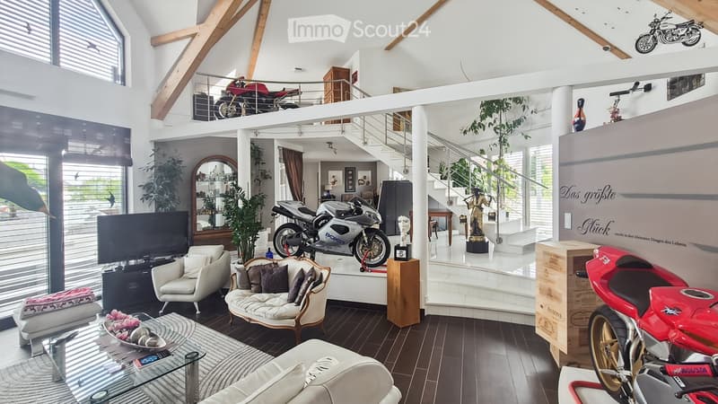 Modern living room with high ceilings, exposed wooden beams, large windows, and a balcony overlooking a staircase area. Features include a white sofa, armchair, coffee table, TV, and several motorcycles displayed on the balcony and in the room.