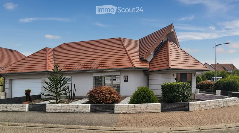 Two-story house with a red-tiled roof, white walls, and a garage. The front yard features neatly trimmed bushes and a small tree, enclosed by a low stone wall.