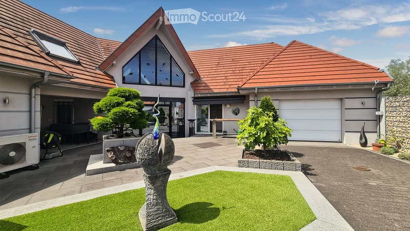 Modern house exterior with a well-maintained garden area, featuring a small lawn, decorative sculptures, trimmed trees, and a paved patio. The house has a distinctive roofline and large windows.