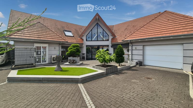 Modern two-story house with a brown tiled roof, white walls, and a spacious driveway. Features include a garage, a small garden with a neatly trimmed lawn, several trees, and a decorative sculpture.