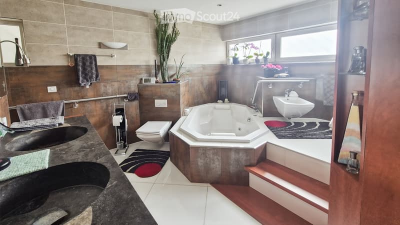 Modern bathroom with a large bathtub, double sink, toilet, and a window with potted plants. Features include a towel rack, a mirror, and decorative rugs.