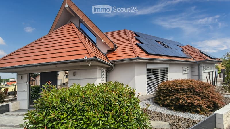 Modern two-story house with red tile roof, solar panels, white walls, and landscaped garden with shrubs and gravel.